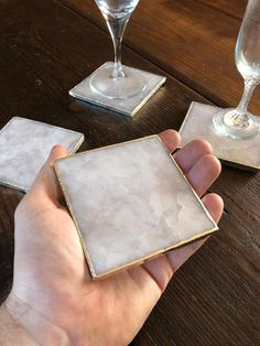 a person is holding some glass coasters on a table with two wine glasses in the background