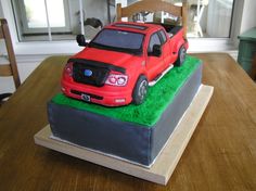 a red truck cake sitting on top of a wooden table next to a green field