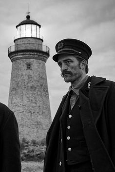 two men standing in front of a light house with one man wearing a hat and coat