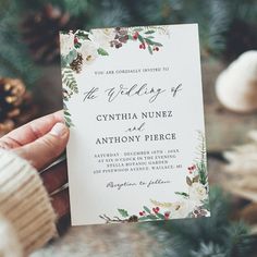 a person holding up a wedding card in front of some pine cones and fir trees