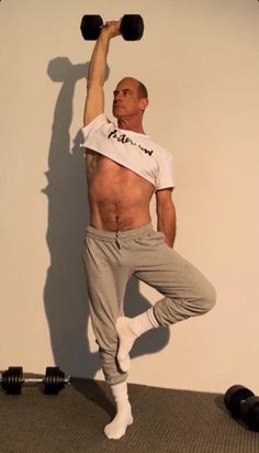 a shirtless man holding two dumbbells while standing on one leg in front of a wall