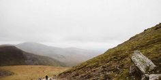 two people walking up the side of a hill on a cloudy day with mountains in the background