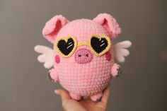 a hand holding a pink crocheted pig with heart shaped glasses on it's face