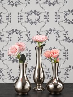 three silver vases with pink flowers in front of a wallpapered background on a table