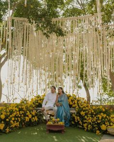 a man and woman sitting on a bench in front of yellow flowers with white strings hanging from the ceiling