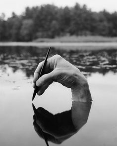 a hand holding a pen writing on top of a body of water with trees in the background