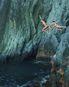 two people jumping off rocks into the ocean from a rocky cliff near a body of water