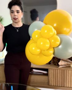 a woman holding balloons in her hand and pointing to the camera with both hands, while standing next to a coffee table