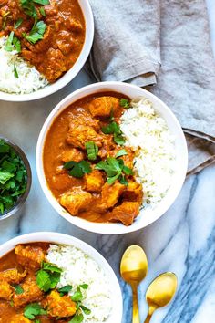 three bowls filled with curry and rice on top of a marble table next to silver spoons