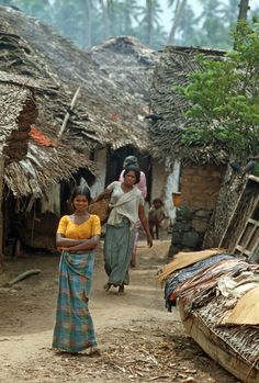 two women walking down a dirt road in front of huts