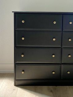 a black dresser with gold knobs on the top and bottom drawers, in front of a white wall