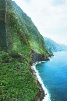 an aerial view of the ocean and mountains near a beach with green grass on both sides