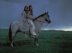 two women riding on the back of a white horse in a green field under a cloudy sky