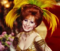 a woman with red hair wearing a gold dress and green feathers on her head, smiling at the camera