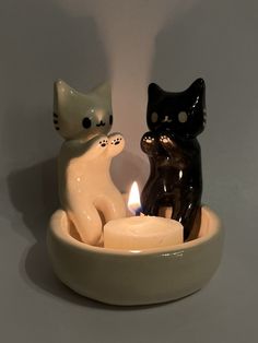 two ceramic cats sitting in a bowl with a lit candle on the side and one cat standing behind it