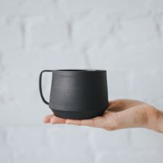 a hand holding a black cup in front of a white brick wall