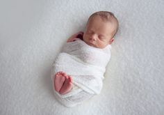 a newborn baby wrapped in a white wrap on top of a white blanket with his eyes closed