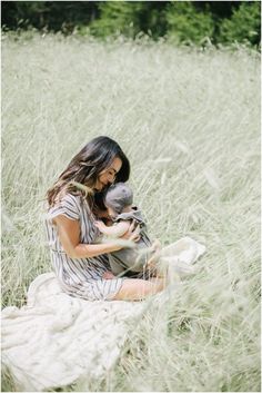 a woman holding a baby in her arms while sitting in tall grass with trees in the background