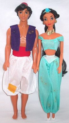 two dolls standing next to each other in front of a white background and one is wearing a blue dress