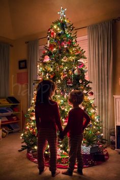 two young children standing in front of a christmas tree with lights on it, looking at each other