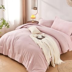 a bed with pink comforter and pillows in a room next to a lamp on a table