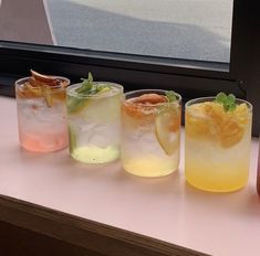 four glasses filled with different types of drinks sitting on a window sill next to an orange slice