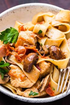 a white bowl filled with pasta and meat on top of a wooden table next to a fork