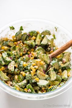 a glass bowl filled with cucumber and corn salad next to a wooden spoon