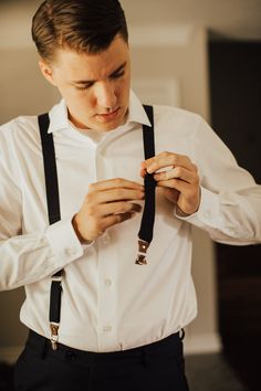 a young man in a white shirt and black suspenders tying his tie