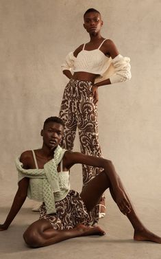 two women pose in front of a wall wearing patterned pants and cropped tops, one with her leg up on the ground