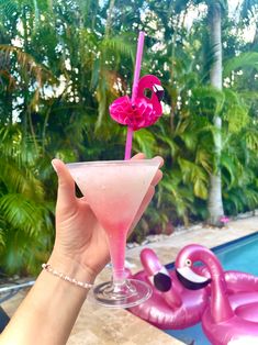 a person holding up a pink cocktail in front of a swimming pool with flamingos