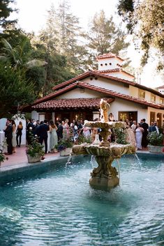 a group of people standing around a fountain in front of a house with a clock on it