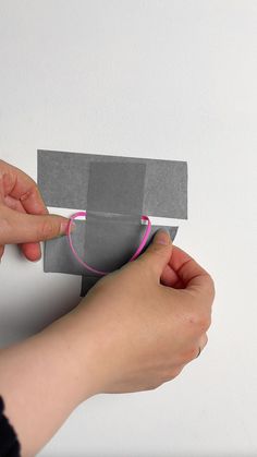 a person is holding a piece of gray paper with a pink ring on it, while another hand holds the edge of an origami board