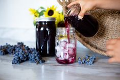 a person pouring red wine into a mason jar filled with ice and blueberries next to sunflowers