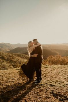 a man and woman standing on top of a grass covered hill hugging in the sun