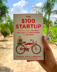 the $ 100 start up book is held in front of a dirt road and palm trees