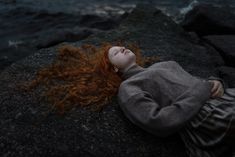 a woman with red hair laying on rocks near the ocean