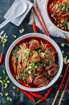 two bowls of beef noodle soup with chopsticks and red peppers on the side