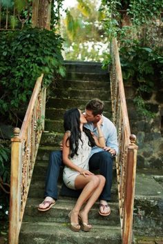 a man and woman kissing on the steps in front of some trees with greenery