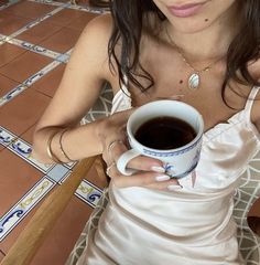 a woman holding a cup of coffee on top of a tiled floor next to a table