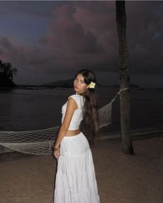 a woman standing on top of a beach next to a hammock with a flower in her hair