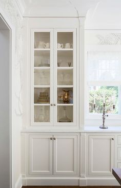 a white china cabinet with glass doors and shelves in the corner, next to a window