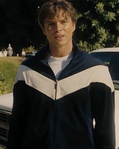 a man standing next to a white car in front of some bushes and trees, wearing a black and white jacket