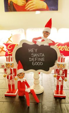 two elfs sitting on top of a table next to a chalkboard with the words hey santa define good written on it