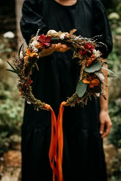 a person holding a wreath with flowers and leaves on it in front of some trees