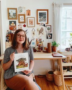 a woman sitting on a chair holding up a book in front of a desk full of pictures