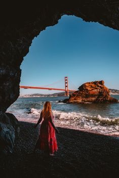 a woman in a red dress is walking on the beach near the water and bridge