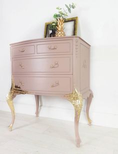 a pink chest of drawers with gold leaf handles and knobs on the top, against a white wall