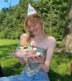 a woman sitting in the grass holding a cake with candles on it and wearing a birthday hat