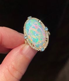 Gorgeous, mesmerizing Ethiopian opal cabochon of AAA quality is framed by dainty diamonds and four blue topazes on each side. The opal sparkles, showcasing streaks and circles of pink, deep blue colors, neon and yellow hues. A truly breathtaking stone! Four soft baby blue round faceted cut topazes add nice detail to the ring. In addition to the frame, diamonds are also placed on the split top part of the band adding more glamour and shine. Very beautiful and elegant ring! Stone: Ethiopian opal, diamonds, blue topazes Stone weight: 7.46 carats Stone shape and size: oval; 12 mm x 22 mm Metal: 14K yellow gold For resizing inquiries, please message directly We gladly accept custom orders using either our designs or design of your choice with stones from our collection or your own.  Please cont Luxury Handmade Yellow Gold Opal Ring, Luxury Ethiopian Opal Engagement Ring, Luxury Adjustable Opal Ring Gift, Luxury Adjustable Opal Ring As Gift, Luxury Gold Opal Ring With Diamond Accents, Luxury Cabochon Opal Ring For Engagement, Luxury Oval Cabochon Opal Ring For Formal Occasions, Opal Rings With Diamond Accents, Oval Shaped, Luxury Opal Ring With Diamond And Gemstone Accents